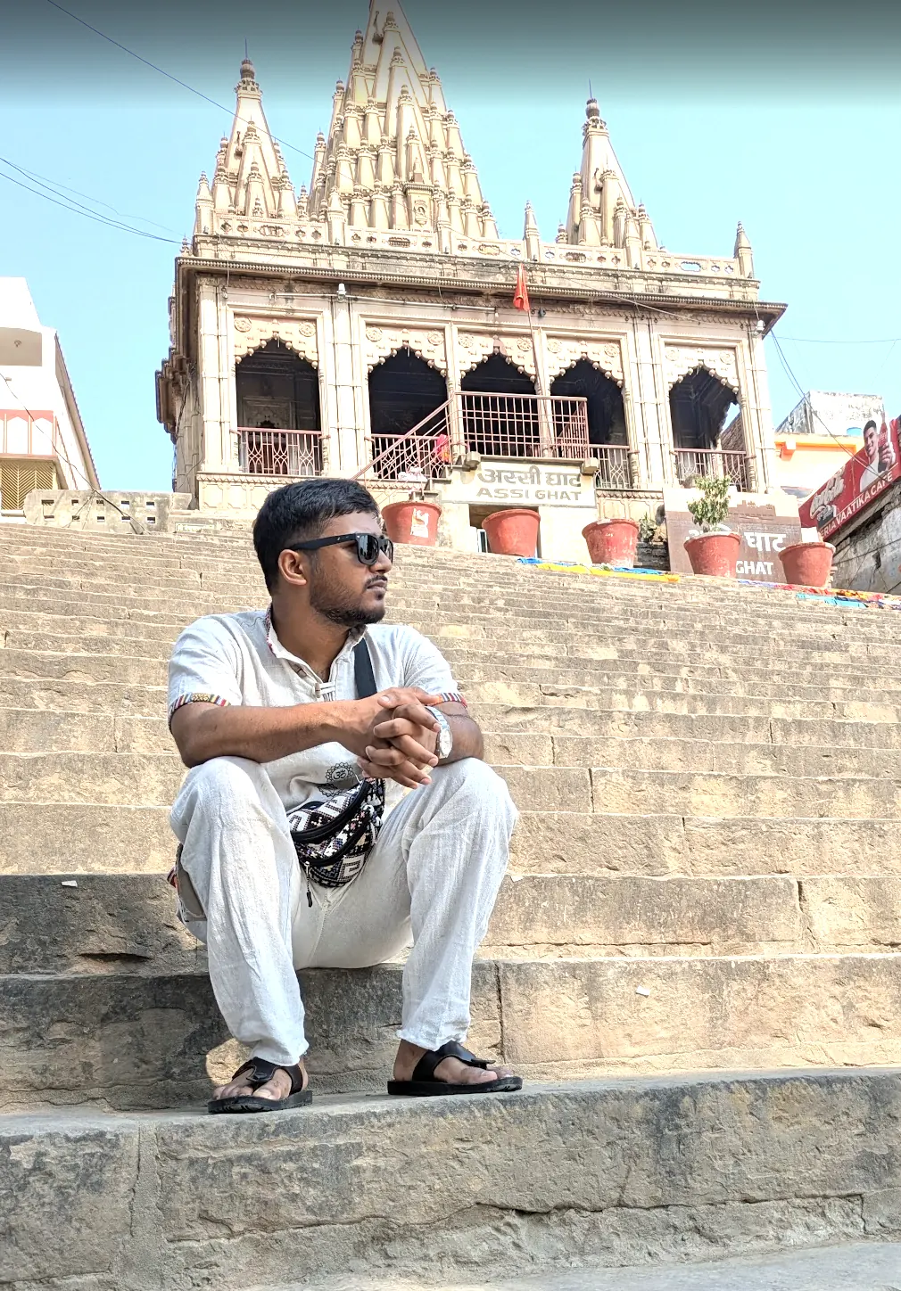 Man in white sitting on the stone steps of a ghat in Banaras, a temple behind him
